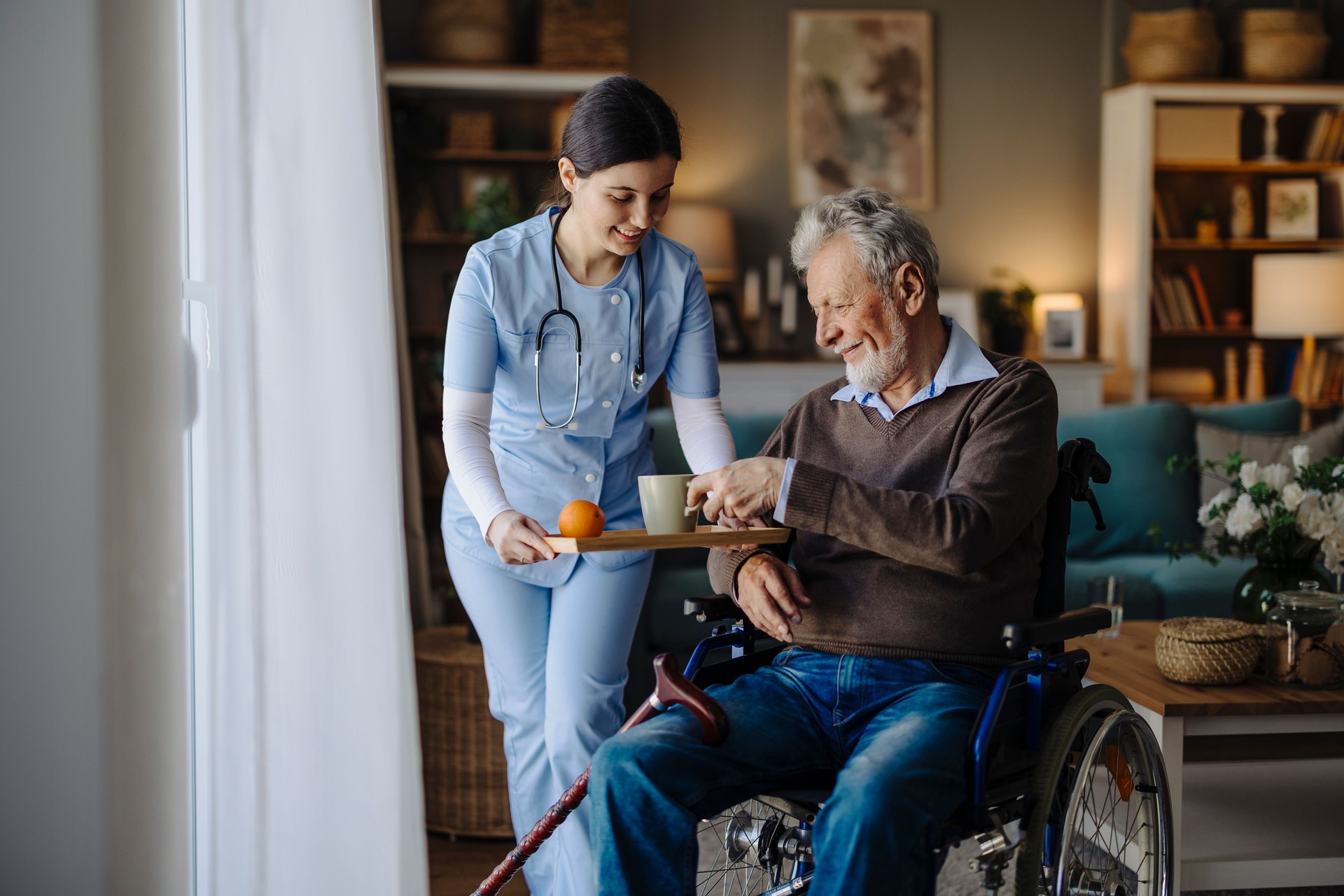 Senior man in wheelchair drinking tea in the nursing home assisted by the caregiver